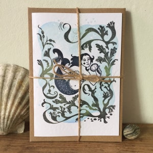 Puede incluir: Una tarjeta con una ilustraci&oacute;n azul y verde de una sirena sosteniendo un espejo. La sirena est&aacute; rodeada de algas marinas y otras plantas acu&aacute;ticas. La tarjeta est&aacute; atada con una cuerda y est&aacute; lista para ser enviada por correo.