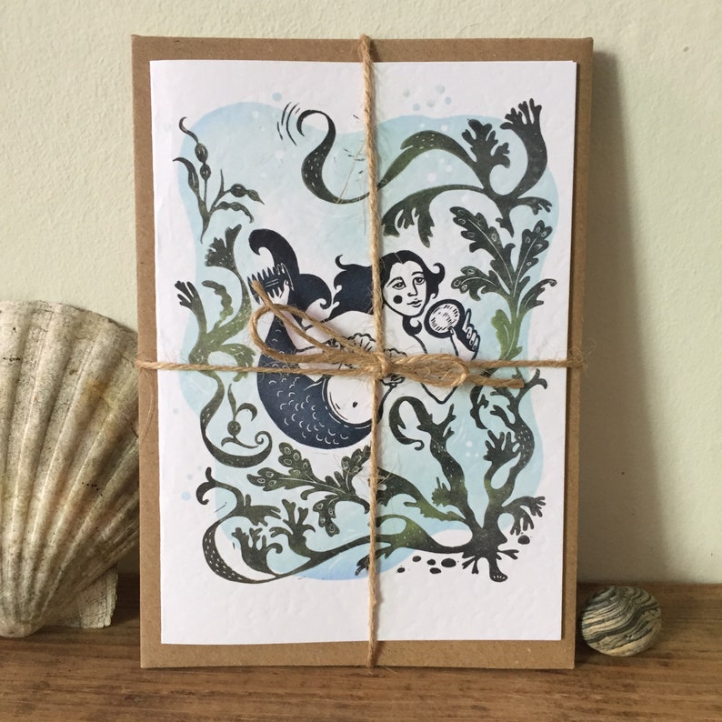 Puede incluir: Una tarjeta con una ilustraci&oacute;n azul y verde de una sirena sosteniendo un espejo. La sirena est&aacute; rodeada de algas marinas y otras plantas acu&aacute;ticas. La tarjeta est&aacute; atada con una cuerda y est&aacute; lista para ser enviada por correo.