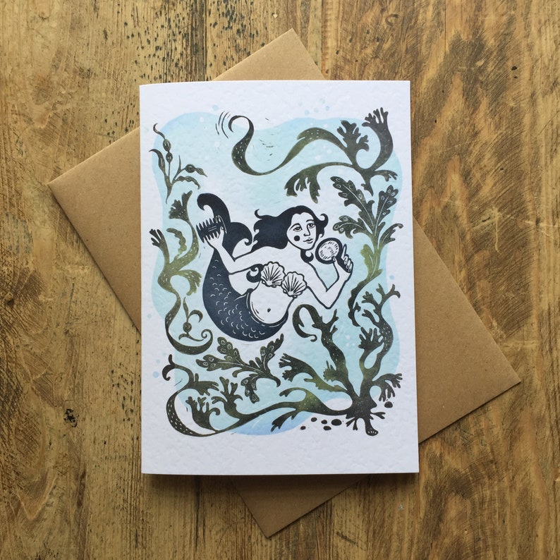 Puede incluir: Una tarjeta de felicitaci&oacute;n con una ilustraci&oacute;n en blanco y negro de una sirena con un sujetador de concha y un peine en el pelo. La sirena est&aacute; rodeada de algas verdes y agua azul.