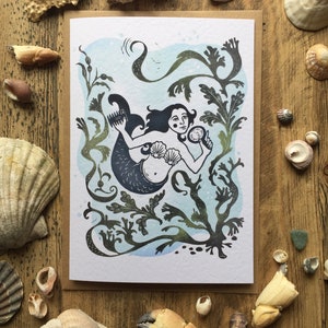 Puede incluir: Una tarjeta de felicitaci&oacute;n con una ilustraci&oacute;n en blanco y negro de una sirena con el pelo largo, sosteniendo un espejo de concha. La sirena est&aacute; rodeada de algas marinas y otras plantas oce&aacute;nicas. El fondo es una acuarela azul claro.