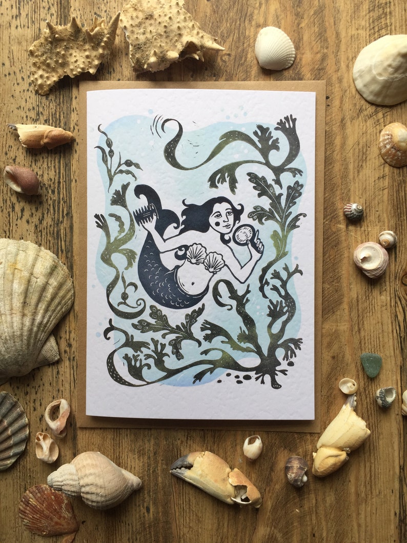 Puede incluir: Una tarjeta de felicitaci&oacute;n con una ilustraci&oacute;n en blanco y negro de una sirena con el pelo largo, sosteniendo un espejo de concha. La sirena est&aacute; rodeada de algas marinas y otras plantas oce&aacute;nicas. El fondo es una acuarela azul claro.