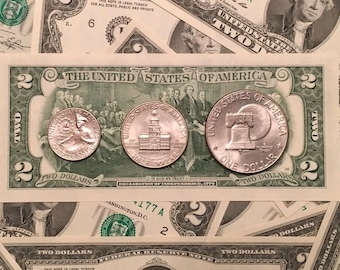 May include: A two dollar bill with three silver coins on top. The coins are a half dollar, a dollar, and a two dollar coin. The coins feature images of the Liberty Bell, Independence Hall, and the Statue of Liberty.