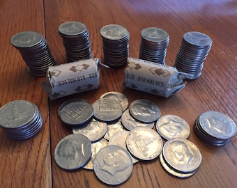 May include: Stacks of silver half-dollar coins and two paper rolls of $10 halves are displayed on a wooden surface. Some coins are scattered, while others are neatly stacked. The coins feature a profile and a building.
