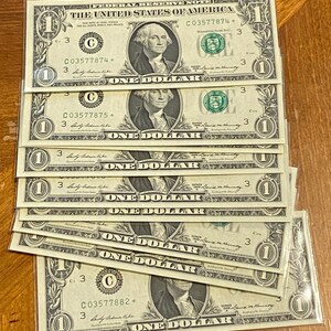 May include: A stack of United States one-dollar bills. Each bill features a portrait of George Washington and the text "ONE DOLLAR". The bills are green and black, with the text "THE UNITED STATES OF AMERICA" at the top.