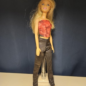 May include: A doll with long blonde hair wearing a red and black patterned tube top and dark gray patterned pants. The doll is standing on a silver stand against a dark blue background.