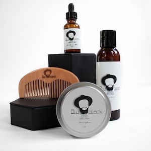 Puede incluir: Un set de cuidado de la barba con un peine de madera, una lata plateada de mantequilla para barba, una botella marrón de aceite para barba y una botella marrón de lavado de barba. Todos los productos están etiquetados con "Olu Black" y presentan una ilustración en blanco y negro de un hombre con barba.