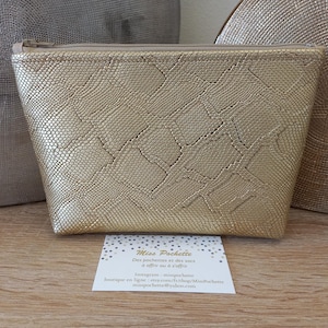May include: A gold-colored, textured cosmetic bag with a zipper closure. The bag has a reptile-like pattern and is displayed on a wooden surface. A business card with text is in front of the bag.