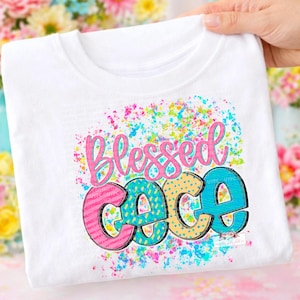 May include: White t-shirt with the words "Blessed Coco" in a colorful, playful design. The word "Blessed" is in pink script, and "Coco" is in blue, pink, and yellow with a splatter effect.