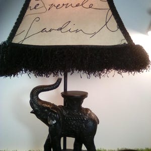 May include: Black table lamp featuring an elephant base. The lamp shade is cream-colored with black script and a black fringe trim. The lamp is on a black base, with a green surface in the foreground.