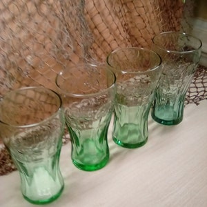 May include: Four green glass Coca-Cola glasses with a vintage design. The glasses are clear with a green tint and have a raised Coca-Cola logo on the front.