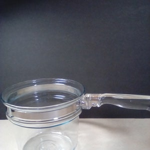 May include: A clear glass double boiler with a silver band and a clear plastic handle.