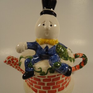 May include: A ceramic teapot shaped like a chimney with a whimsical character sitting on top. The character is a white egg with a black top hat, blue suit, and yellow bow tie. The teapot is decorated with red brick, green vines, and white flowers.