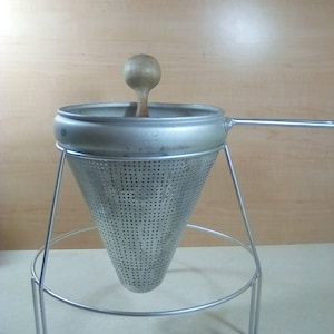 May include: A silver conical strainer with a wooden handle sits on a metal stand. The strainer has a fine mesh and is designed for straining liquids.