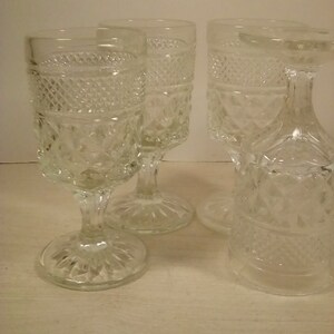 May include: Four clear, vintage-style stemmed glasses. The glasses have a diamond pattern on the bowl and a decorative base. One glass is upside down. The glasses are on a light-colored surface.