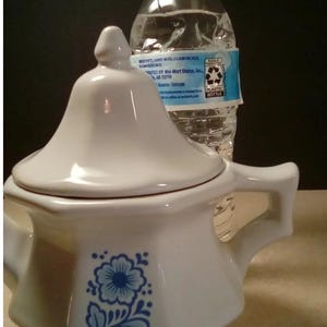 May include: A white ceramic sugar bowl with a lid and two handles, featuring a blue floral pattern on the front. The bowl has a hexagonal shape and is positioned in front of a clear plastic water bottle with a white cap and a blue and white label.
