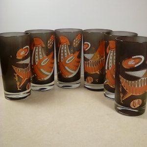 May include: A set of six cylindrical drinking glasses made of dark smoke-colored glass. Each glass features a repeating graphic print in orange, white, and black, depicting stylized bird-like figures and geometric shapes, with the signature Georges Briard.