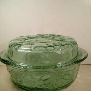 May include: A vintage green glass serving dish with a lid. The dish has a round shape with embossed floral patterns on the sides and lid. The lid features a textured design. The dish is made of transparent green glass.