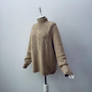 Puede incluir: Un suéter beige de punto trenzado con cuello alto. La prenda se exhibe en un maniquí sobre un fondo blanco. El diseño de punto trenzado está centrado en la parte delantera. Las mangas son largas y el material parece ser de punto de peso medio.