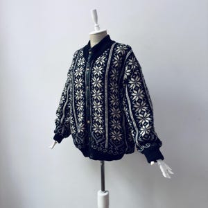May include: A black and white patterned cardigan with a snowflake design. The cardigan has a button-up front, a black collar, and black cuffs. The garment is displayed on a mannequin.