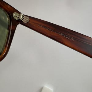 May include: Close-up of a pair of sunglasses with a brown tortoiseshell frame. The hinge is silver, and the arm of the glasses has the text "B&L RAY-BAN U.S.A." printed on it. The lens is green.
