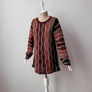 Op de afbeelding: Een trui met lange mouwen in tinten bordeaux, bruin en oranje, weergegeven op een mannequin. De trui heeft verticale golvende strepen met een crèmekleurig stikseldetail. De halslijn is diep bordeaux.