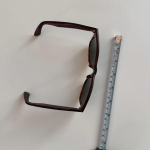 May include: Brown tortoiseshell sunglasses with dark lenses. The sunglasses are measured with a tape measure, showing a length of approximately 6 inches. The frames have a classic, rectangular shape.
