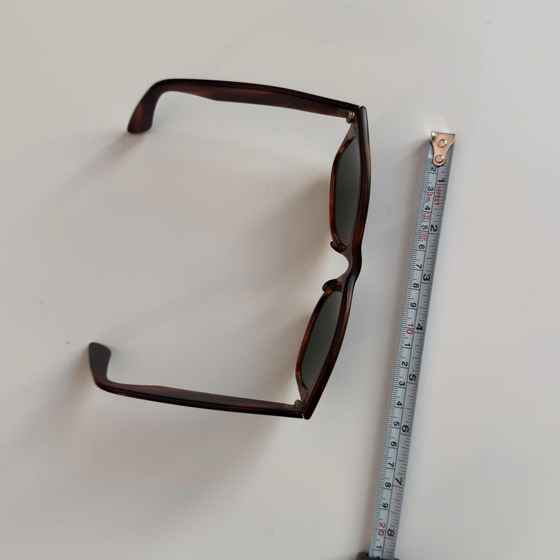 May include: Brown tortoiseshell sunglasses with dark lenses. The sunglasses are measured with a tape measure, showing a length of approximately 6 inches. The frames have a classic, rectangular shape.