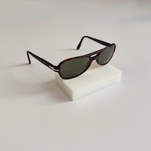 May include: Aviator-style sunglasses with a tortoiseshell frame and dark green lenses. The sunglasses are resting on a white rectangular block, showcasing their classic design. The frame features a silver accent near the hinges.