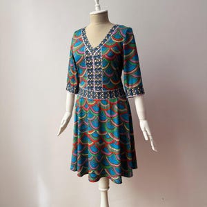 Op de afbeelding: Een kleurrijke jurk met een V-hals en driekwartmouwen. De jurk heeft een levendig ontwerp van overlappende, schubachtige vormen in tinten blauw, groen, rood en roze. Witte biezen accentueren de halslijn, mouwen en taille.