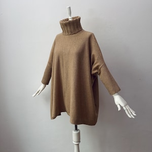 May include: A tan-colored, oversized turtleneck sweater displayed on a mannequin. The sweater features a ribbed turtleneck, long sleeves with ribbed cuffs, and a loose, flowing body. The garment is made of a soft, knitted material.