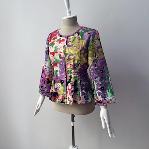 Può includere: Giacca con stampa floreale con scollo tondo e maniche svasate. La giacca presenta un mix di colori, tra cui viola, giallo, verde e rosso. L'indumento è esposto su un manichino.