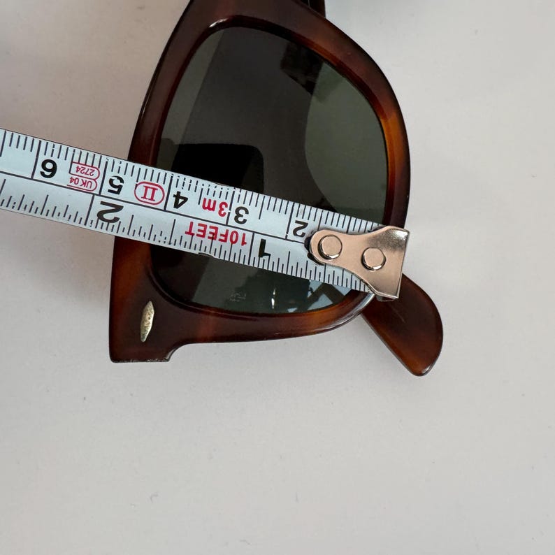 May include: Brown sunglasses with dark lenses are measured with a tape measure. The tape measure shows a measurement of approximately 3 inches. The sunglasses have a classic, rectangular frame.