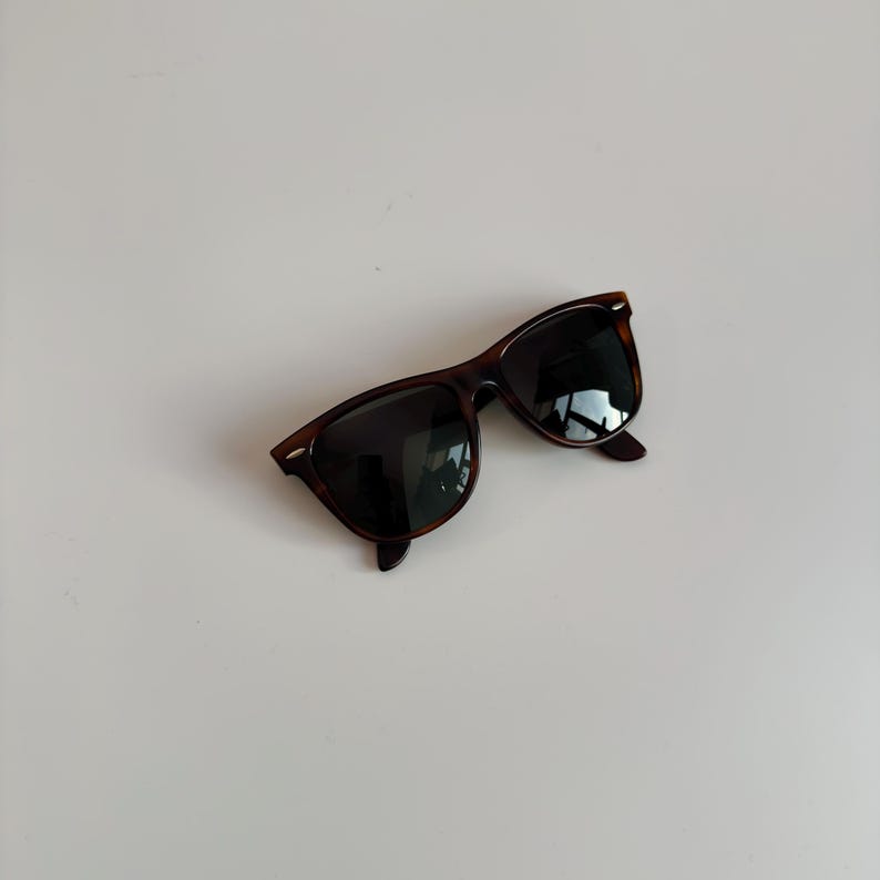 May include: Sunglasses with a classic design. The frames are a dark brown tortoiseshell pattern, and the lenses are black. The sunglasses are angled slightly, showing the shape of the frames and lenses.