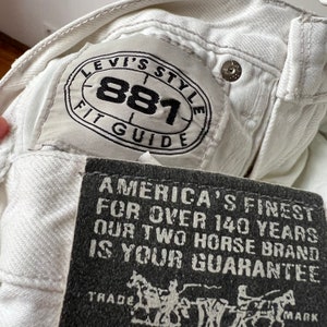 May include: Close-up of white denim jeans with a Levi's Style 881 Fit Guide label. A black tag reads "America's Finest For Over 140 Years Our Two Horse Brand Is Your Guarantee". The jeans have a button closure.