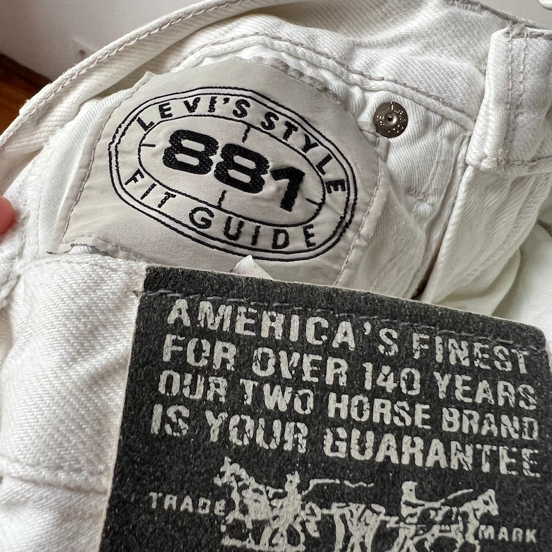 May include: Close-up of white denim jeans with a Levi's Style 881 Fit Guide label. A black tag reads "America's Finest For Over 140 Years Our Two Horse Brand Is Your Guarantee". The jeans have a button closure.