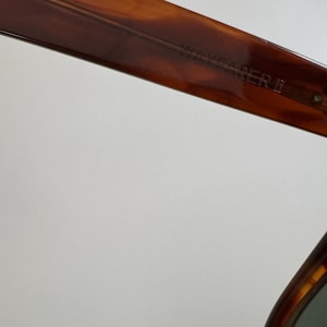 May include: Close-up of the upper frame of Wayfarer II sunglasses. The frame is a tortoiseshell pattern with a dark brown color. The text "WAYFARER II" is visible on the frame.