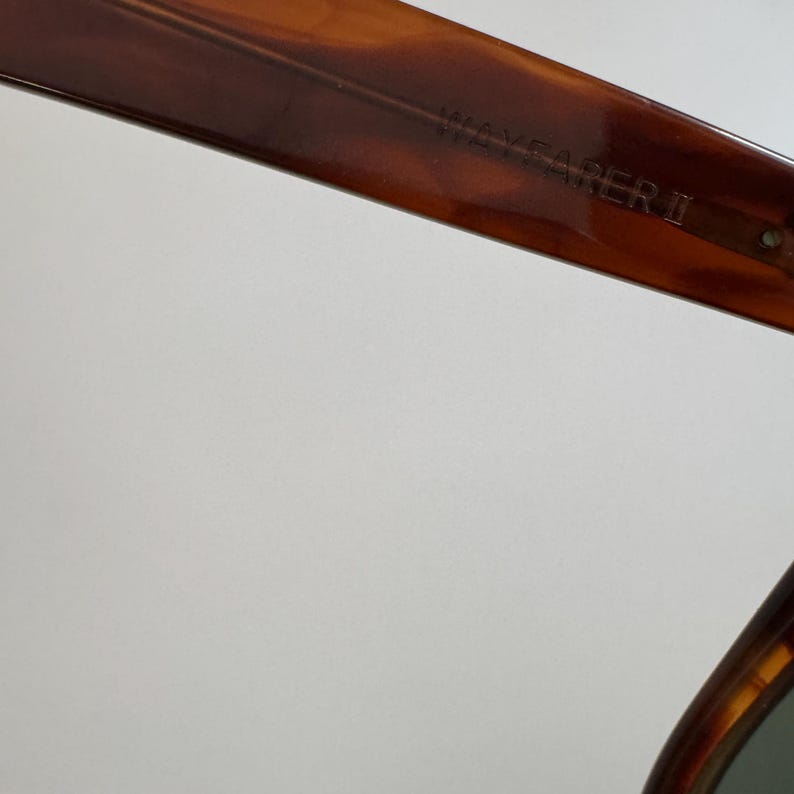 May include: Close-up of the upper frame of Wayfarer II sunglasses. The frame is a tortoiseshell pattern with a dark brown color. The text "WAYFARER II" is visible on the frame.