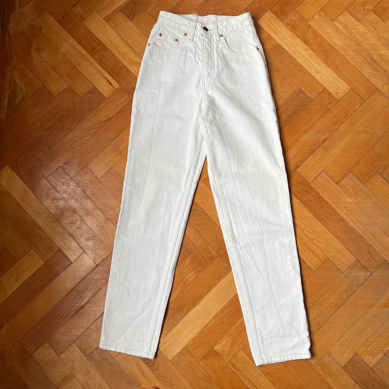 May include: White denim jeans with a high waist and straight leg. The jeans feature a button and zip fastening, classic five-pocket design, and copper rivets. White stitching is visible. The jeans are laid flat on a wooden surface.