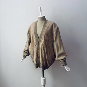 May include: A tan leather cardigan featuring a V-neckline, long sleeves with ribbed cuffs, and a ribbed hem. The garment includes a front button closure with four visible buttons and two side pockets, constructed with multiple leather panels.