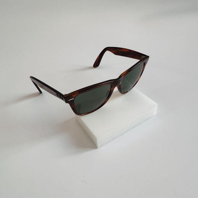 May include: Brown tortoiseshell sunglasses with dark green lenses. The sunglasses are resting on a white rectangular block. The frames have a classic, retro design.