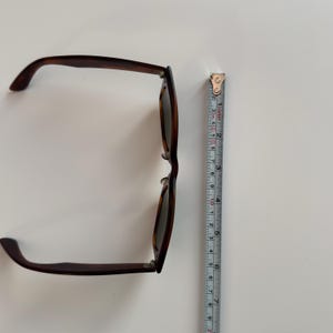 May include: Brown tortoiseshell sunglasses with dark lenses. The sunglasses are next to a measuring tape. The sunglasses are approximately 6 inches long.