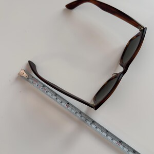 May include: Tortoiseshell sunglasses with dark lenses. The sunglasses are brown and black with a classic frame design. A measuring tape is placed next to the sunglasses, showing a length of approximately 8 inches.