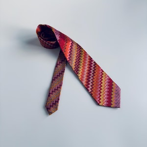 Puede incluir: Una corbata colorida con un patrón en zigzag en tonos de rojo, naranja, amarillo y morado. La corbata está hecha de una tela texturizada y se muestra sobre una superficie blanca. Es un accesorio de moda.