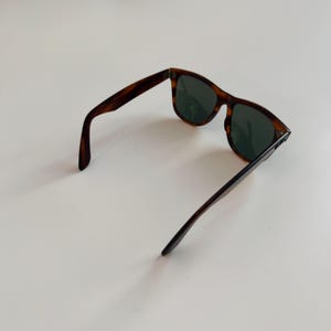 May include: Tortoiseshell sunglasses with dark green lenses. The sunglasses have a classic rectangular frame and are open, displaying the arms and lenses. The frame has a brown and black pattern.