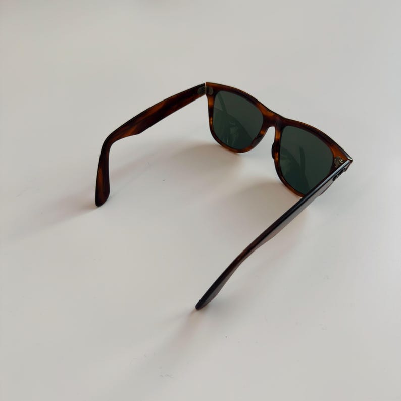 May include: Tortoiseshell sunglasses with dark green lenses. The sunglasses have a classic rectangular frame and are open, displaying the arms and lenses. The frame has a brown and black pattern.
