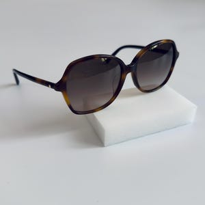 Puede incluir: Gafas de sol carey con lentes oscuras. Las gafas de sol tienen una montura redondeada y de gran tamaño y varillas oscuras. Las gafas están sobre una superficie blanca. Las lentes tienen un tinte degradado.