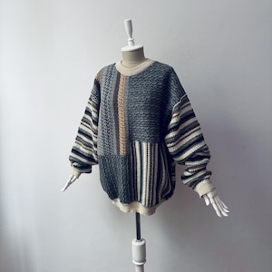 Puede incluir: Un suéter de manga larga con cuello redondo que presenta un diseño de parches con paneles gris oscuro texturizados, rayas verticales en tonos marrón, crema y azul, y un cuello, puños y dobladillo acanalados en color crema.