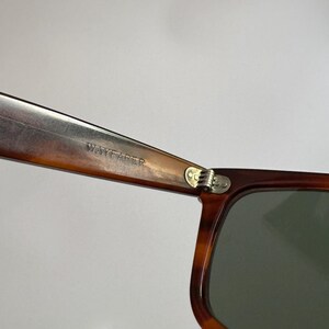 May include: Close-up of Wayfarer sunglasses with a brown tortoiseshell frame and dark green lenses. The text "WAYFARER" is visible on the arm of the sunglasses. The hinge is silver.