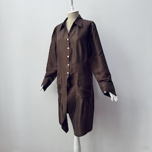 Può includere: Un lungo abito camicia marrone con maniche lunghe e due tasche anteriori. L'abito è esposto su un manichino con sfondo bianco. L'abito ha un colletto e bottoni bianchi.