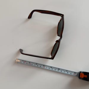 May include: Brown tortoiseshell sunglasses with dark lenses. A measuring tape is placed next to the sunglasses. The sunglasses measure approximately 6 inches in length.
